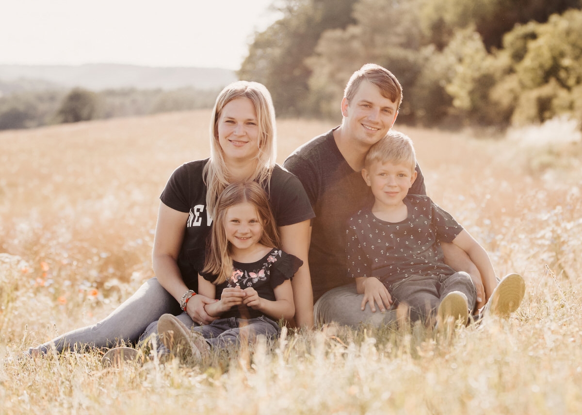 Eine Familie mit zwei Kindern sitzen gemeinsam in einem Feld. Alle gucken direkt den Betrachter an und lächeln. Fotografin: Claudia Nürnberger