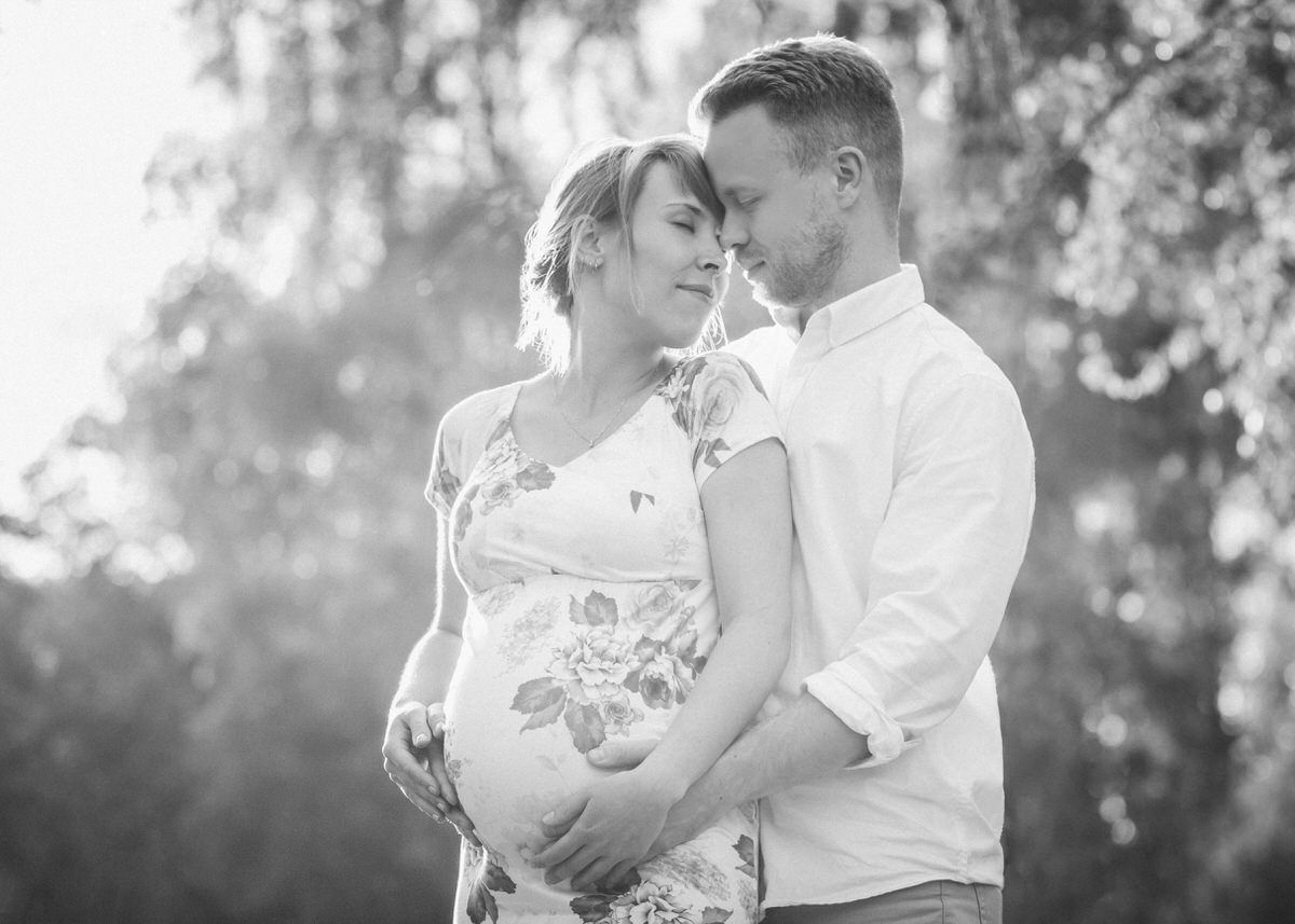 Mann steht hinter seiner schwangeren Frau. Ihre Hände liegen aufeinander auf ihrem Babybauch während sie ihm verliebt ihr Gesicht zuwendet. Beide halten die Augen geschlossen in diesem zärtlichen Moment.
 Fotografin: Claudia Nürnberger